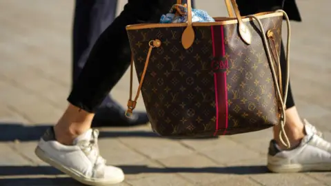 Getty Images A guest wears black suede pants, a brown LV monogram in coated canvas Neverfull handbag from Louis Vuitton, white leather sneakers, outside Louis Vuitton, during Paris Fashion Week - Womenswear Spring/Summer 2023, on October 04, 2022 in Paris, France.