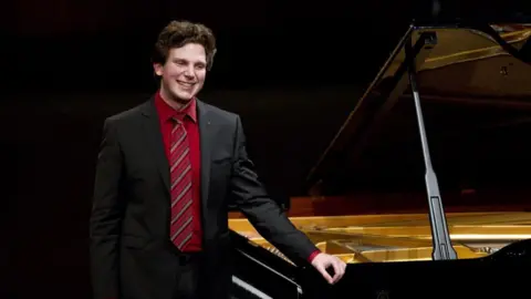 Getty Images British-Australian pianist Jayson Gillham wearing a suit stands beside a grand piano.
