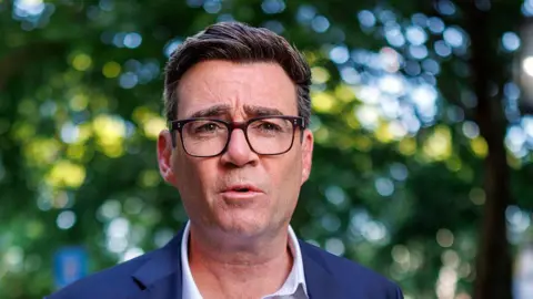 Getty Images Andy Burnham wearing a white shirt and blue blazer. He is looking at the camera, and is wearing glasses.