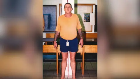 Supplied: NT Police A tall man in a yellow t-shirt and blue short stands against a measuring tape in an office