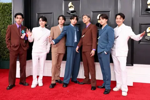 Amy Sussman / Getty Images BTS stand in a line while posing wearing suits with the Grammys logo in the bacgkround, at the 2022 Grammys red carpet