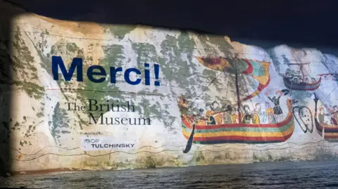 British Museum White cliffs of Dover at night, covered in a giant projection with images of wooden boats from the tapestry, and "Merci"