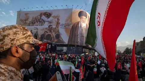 NurPhoto via Getty Images an Iranian security officer amid crowds in front of a billboard showing the late supreme leader at his funeral