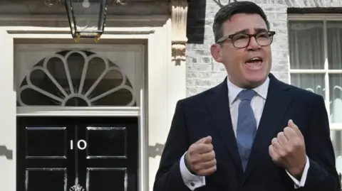 Jeff Overs/BBC Andy Burnham is wearing a dark-coloured suit jacket, a white shirt and blue tie. He is also wearing glasses. He has black-coloured short hair. He is stood in Downing Street making a speech. Behind him is a black door with the number 10 on it.