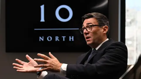 BBC Andy Burnham, wearing a dark suit, gestures with his hands while speaking during an interview. Behind him is a TV with the logo for No 10 North.