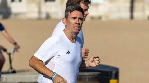 Tayfun Salci Andy Burnham wearing a white Adidas t shirt and black athletic shorts, running next to Green Park. The sun is on his face