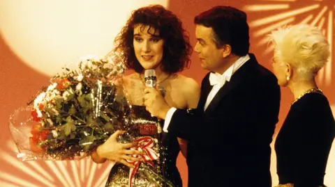 Getty Images A young Celine Dion holding flowers as she is interviewed a the Eurovision Song Contest. She is wearing a glittery strapless dress