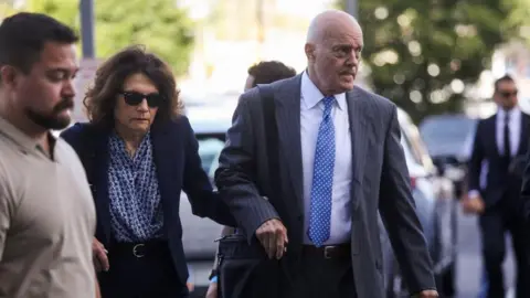 Reuters A balding man in a grey suit holds the hand of a woman in a navy blue suit who is wearing sunglasses as both are walking