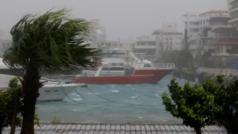 Reuters High winds and wet weather at a marina in Ishigaki Island, in Japan's Okinawa Prefecture. Wind blows through a palm tree and boats are seen on a choppy sea close to the quay.