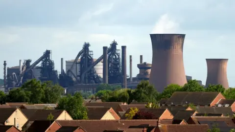 PA Media Brown roofs and leafy green trees sit in front of the squat brown blast furnace smokestacks and industrial pipes and silos of the British Steel plant in Scunthorpe.