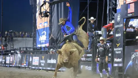 BULL STOCK MEDIA A professional bull rider, wearing a cowboy hat and blue shirt featuring the triangular logo of US Space Force, clings on while riding on the back of a massive, brown horned bull in a stadium filled with dirt 