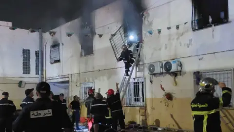 Algeria's General Directorate for Civil Protection A fireman up a ladder removing a grill from a window from which black smoke is billowing. He is gesturing to other fire officials below who are pointing - some wearing red hard hats.
