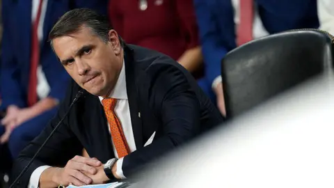 Getty Images Todd Blanche wears a balck suit with an orange tie. He is seen sitting at a desk at a Senate confirmation hearing, with the frame tilted towards the left so the image appears at an angle.