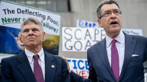 Bloomberg via Getty Images Former Olympian David Hearn, left, and attorney Norman Eisen, both in blue suits, white shirts and purple ties, stand in front of signs in support of Hears. One reads "Scapegoat" 
