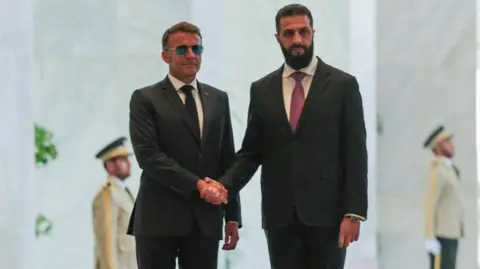 Reuters Macron wears a pair of sunglasses as he shakes hands with al-Sharaa