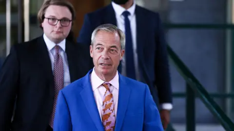 Reuters Nigel Farage walking outside, wearing a blue suit with a pink tie, decorated with a fish pattern. There are two men behind him, both wearing dark suits. 