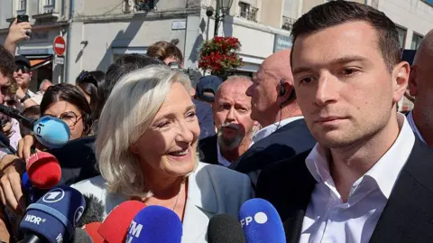 Getty Images A woman stands in a crowd smiling in front of microphones while a man to her right stands beside her with no expression