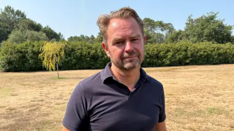 A man dressed in a dark blue polo shirt top stands in the middle of lawn that has been left brown and dry by the heat.