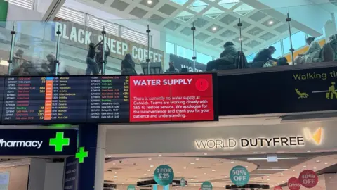 Supplied A red sign alerts passengers to the water supply issue on a screen in a terminal building. A World Duty Free sign is underneath and a pharmacy sign is nearby. A further screen shows flight information.