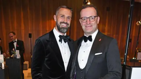 Tristar Media/Getty Images Jens Spahn (R) in glasses, a grey jacket and white shirt and bowtie beside his husband Daniel Funke who has as beard and a black velvet jacket with a white shirt and bowtie