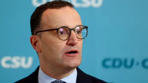 Reuters Jens Spahn speaks with German media during a party meeting, he is wearing a black suit and black glasses.
