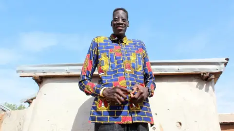 BBC A smiling Sulemana Abdul Samed, also known as Awuche, stands in front a a building, towering over its roof. He wears a colourful shirt. 