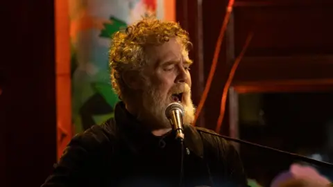 PA Media Glen Hansard has curly reddish/grey hair and beard and is singing while playing a guitar