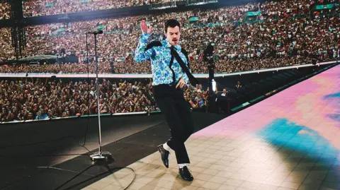 Shuttershock Harry Styles dancing on stage at Wembley wearing a blue floral shirt, black tie, black trousers, white socks and black patent shoes. Thousands of people can be seen standing or watching him from the stands