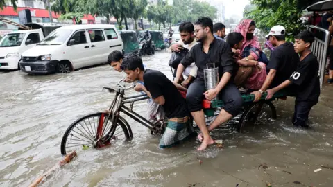 Reuters Several people push a heavily laded tricycle through floodwaters in Dhaka