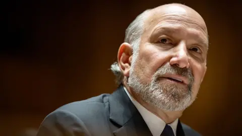Getty Images Close-up portrait of Howard Lutnick with a short grey beard and receding hairline, wearing a dark suit jacket, white shirt, and dark tie. The person is turned slightly to the side and looking toward the camera. The background is softly blurred with warm brown and amber tones, creating a shallow depth-of-field effect.