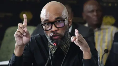 Gallo Images via Getty Images Vusimusi "Cat" Matlala, with the collar of a Fendi shirt sticking out above his jumper, gesticulates as he talks into a microphone.