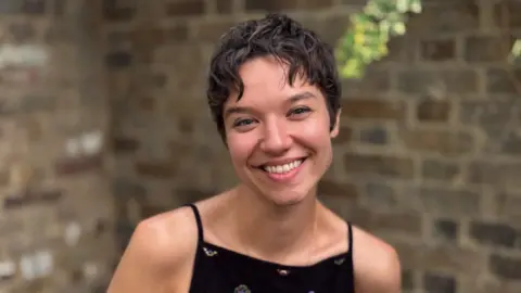 Layla Kornota The image shows a medium close-up portrait of a woman short, dark brown hair.  She is smiling broadly, showing her teeth, and looking directly toward the camera. She is wearing a black top with small, scattered embroidered patterns.  