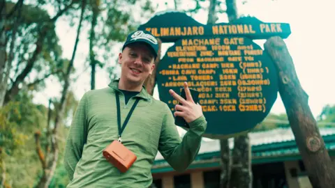 Aaron Fitzmaurice Aitch poses in front of a Kilimanjaro National Park sign. He is wearing a green long-sleeved jumper and a baseball cap. 

