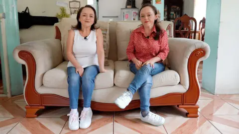 JORGE PEREZ / BBC María Luisa sits on a cream sofa with her sister María del Cisne. María Luisa has shoulder-length brown hair and is smiling, wearing a white vest, jeans and trainers. María del Cisne has longer dark hair, with the front pinned back, a red shirt with white polka dots and is also wearing jeans and white trainers.
