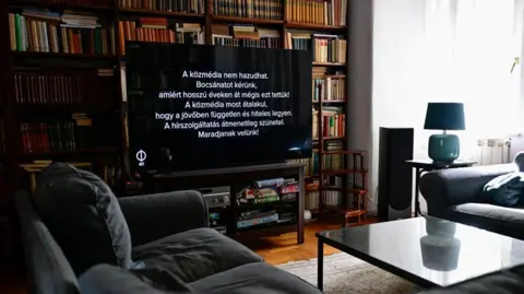 Reuters A TV screen is seen inside a living room in front of a large bookcase and behind a coffee table and two sofas. The screen is black with white writing that reads "State media cannot lie. We apologise for having done so for years".