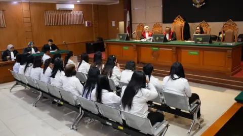 BBC Indonesian Two rows of women wearing white tops and black trousers sit in front of a judge in a courtroom