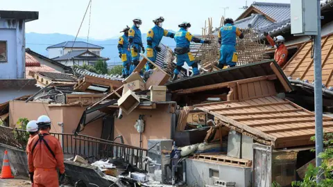 JIJI Press / AFP via Getty Images Rescue workers in overalls stand on top of a collapsed house