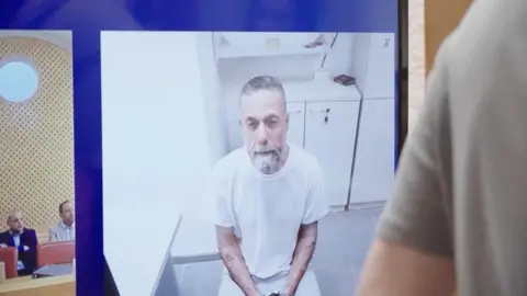 Reuters Detained Palestinian medic Dr Hassam Abu Safiya is pictured on a screen wearing a white shirt in prison during an Israeli Supreme Court hearing on 10 June 2026