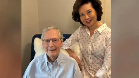 Office of Mitch McConnell Mitch McConnell sits in a chair and smiles at the camera. His wife Elaine Chao sits next to him and also smiles at the camera. 