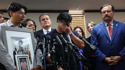 Reuters A man holds his hands over his eyes as he stands before a bank of microphones and a man holding up pictures of Lorenzo Salgado Araujo