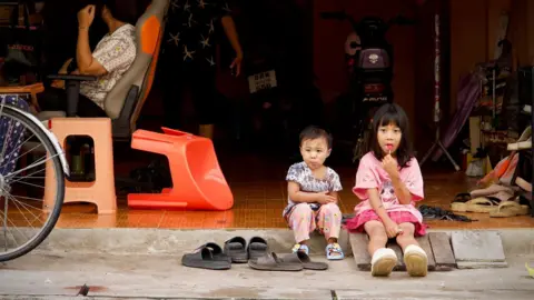 BBC/ Lulu Luo A young girl, lollipop in hand, sitting on the floor with another young child. 