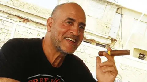 US Department of Justice Daniel Siad, a man with a shaved head and stubble, wearing a black T-shirt, sits outside in what appears to be a sunny street, smiling with a large cigar raised in his hand.