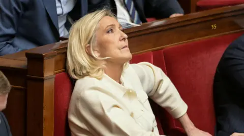 Getty Images Marine Le Pen sits in France's National Assembly in a red velvet seat. She is biting her lip and looking up. She wears a cream suit. 