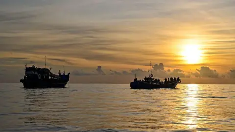 AFP via Getty Images Two boats carrying lots of people float in a sea against a setting sun