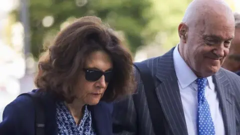 Reuters Charlie Kirk's mother, wearing sunglasses and a blue blazer over a blue patterned blouse, arrives at the Utah County courthouse hand-in-hand with her husband, wearing a striped suit jacket and polka dot blue tie