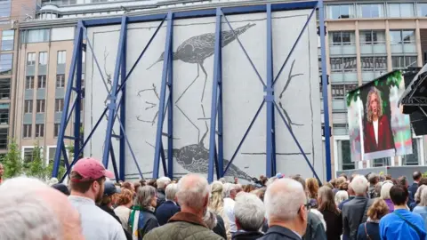 EPA A large crowd stand in front of an artwork depicting a bird reflected in water. The large artwork is covered by a blue steel frame.
