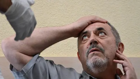 Alexander NEMENOV/AFP via Getty Images A man in a grey shirt holds his right arm to his forehead as he looks at a man out of the picture who is wearing a medical glove