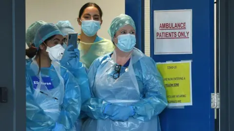 Getty Images Four medics are standing at the doors of an emergency department in the pandemic. They are wearing plastic blue gowns and have face masks on. One has a sign on her chest saying "ED Nurse". 