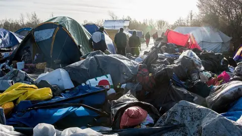 Getty Images A makeshift camp of tents and tarpaulins stands in an open area during daylight, with piles of rubbish and discarded belongings in the foreground. Several people walk between the tents along a muddy path, and low sunlight casts long shadows across the scene.
