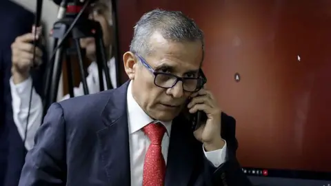 AFP via Getty Images Ollanta Humala speaks on the phone as he leaves court in 2025. He wears a navy suit and red tie